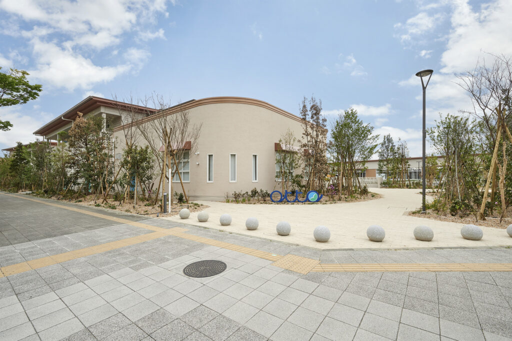 曲線を描く屋根の建物と植栽帯。奥にロゴサインが見える施設の外構