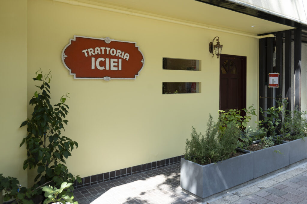 TRATTORIA ICIEIの店舗外観。淡い黄色の壁面に木枠の看板が掛かり、アプローチには小口タイルと植栽が並ぶ