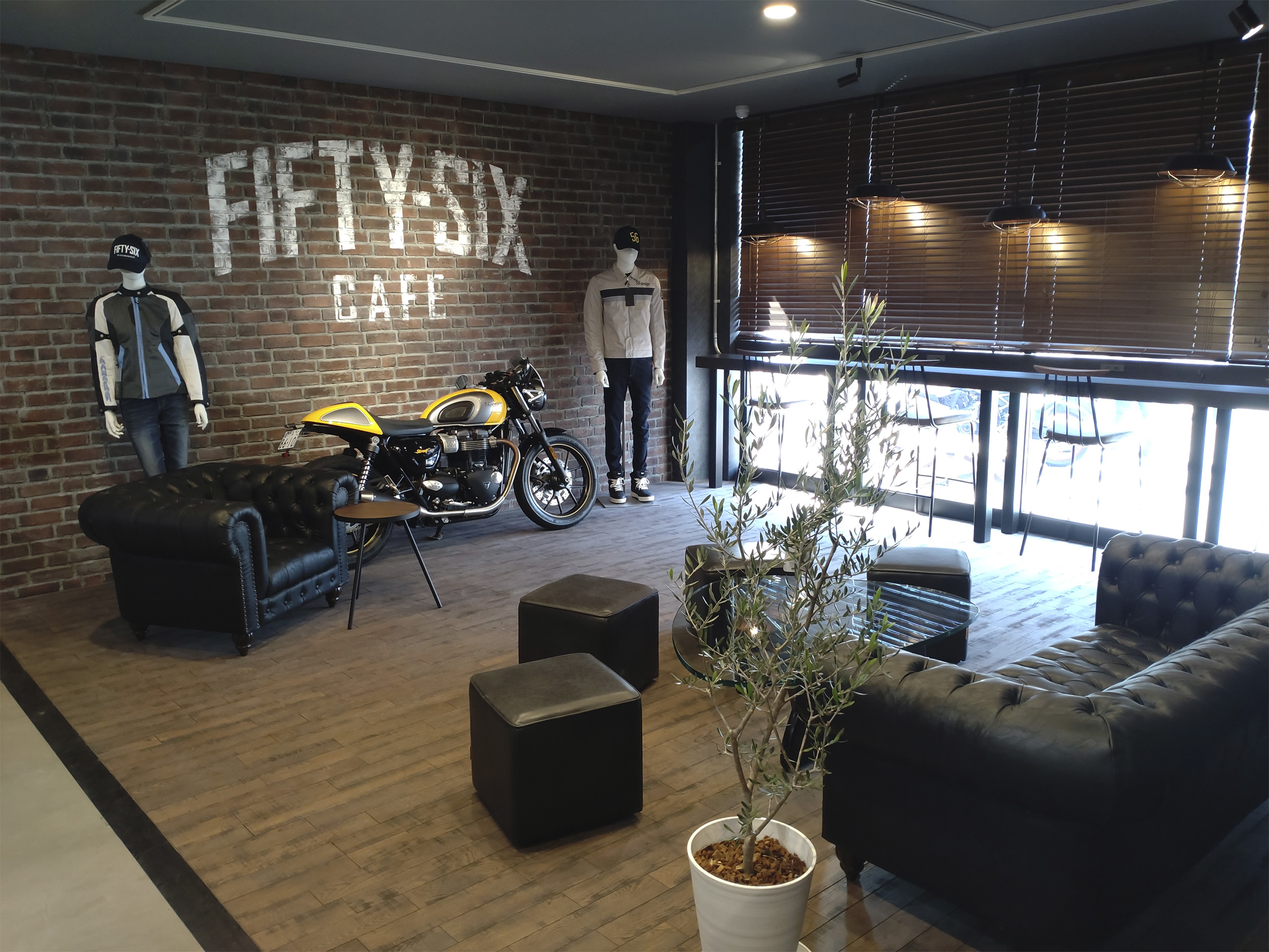 56design NARA operated by RICOLAND のカフェスペース。キャンブリック STANDARD STD-30 のレンガ調タイルの壁にロゴがあり、レザーソファとバイクが並ぶ